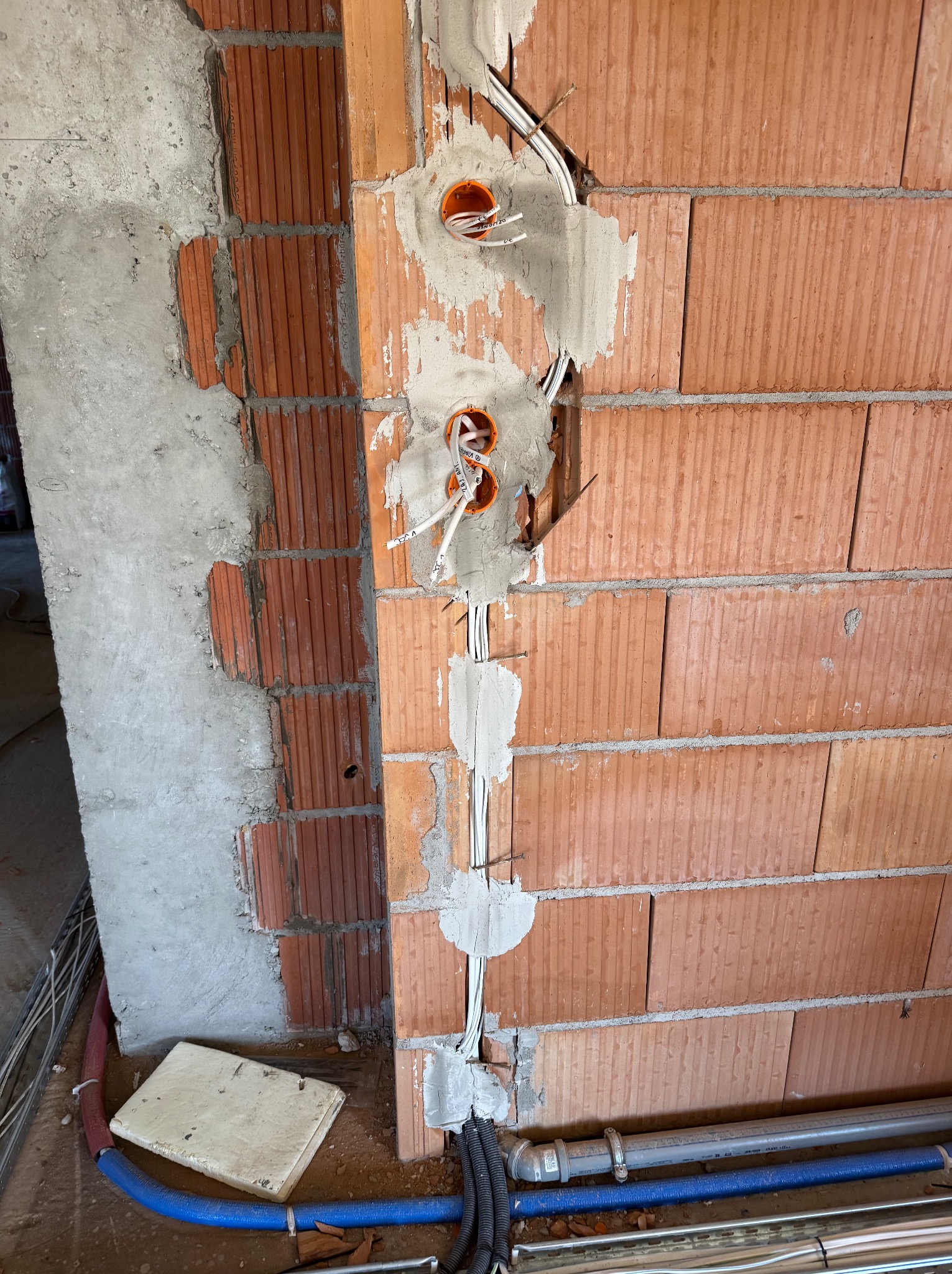 Świeżo wybruzdowana ściana z czerwonej cegły z widocznymi puszkami i przewodami elektrycznymi, zabezpieczonymi gipsem, Kędzierzyn-Koźle. Widoczne także rury instalacyjne przy podłodze.