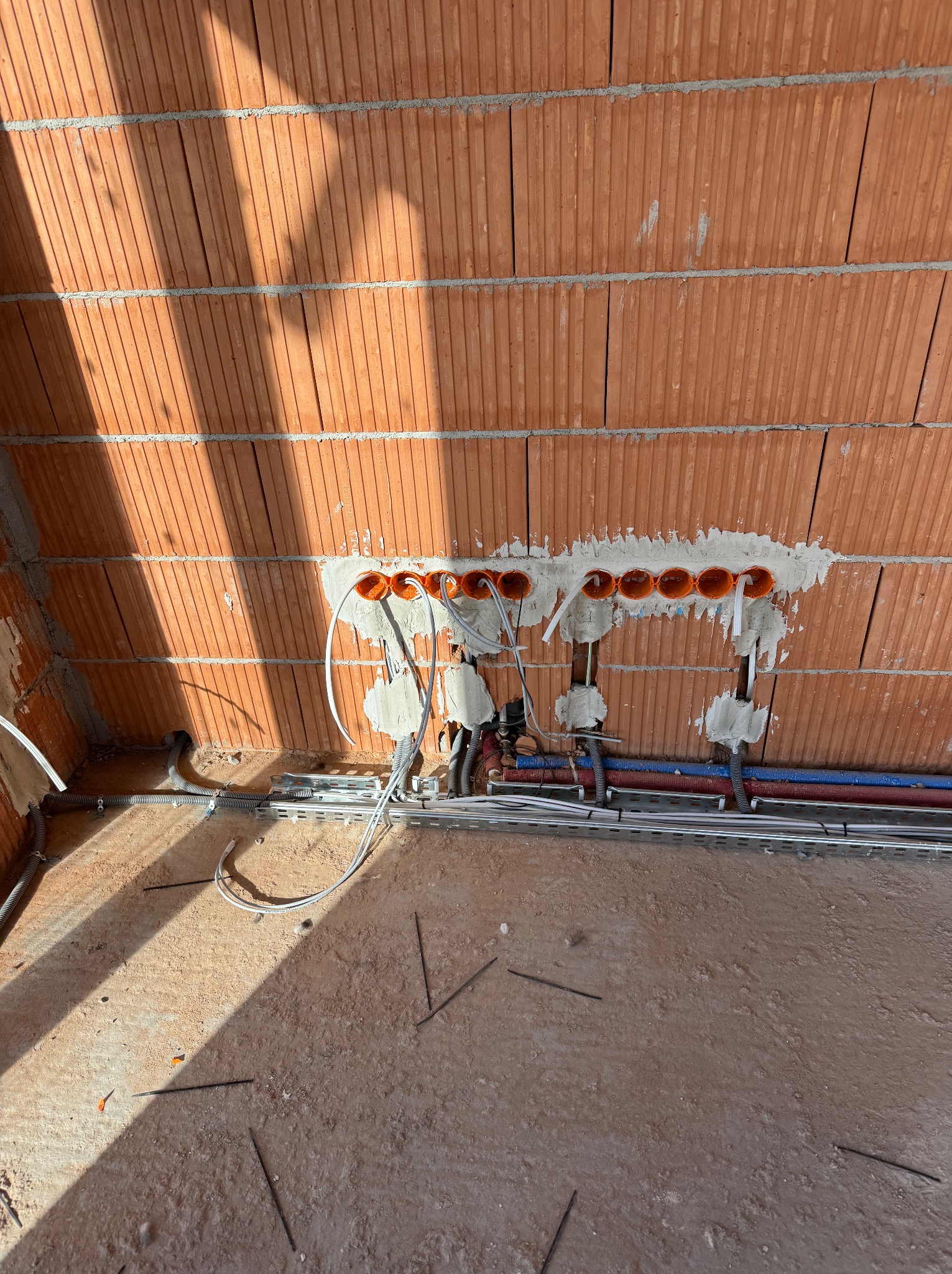 Surowa ściana z cegły z zamontowanymi pomarańczowymi puszkami pod gniazdka, częściowo zamurowanymi i z wyprowadzonymi przewodami; widoczne rury instalacyjne i metalowe korytka kablowe na podłodze...