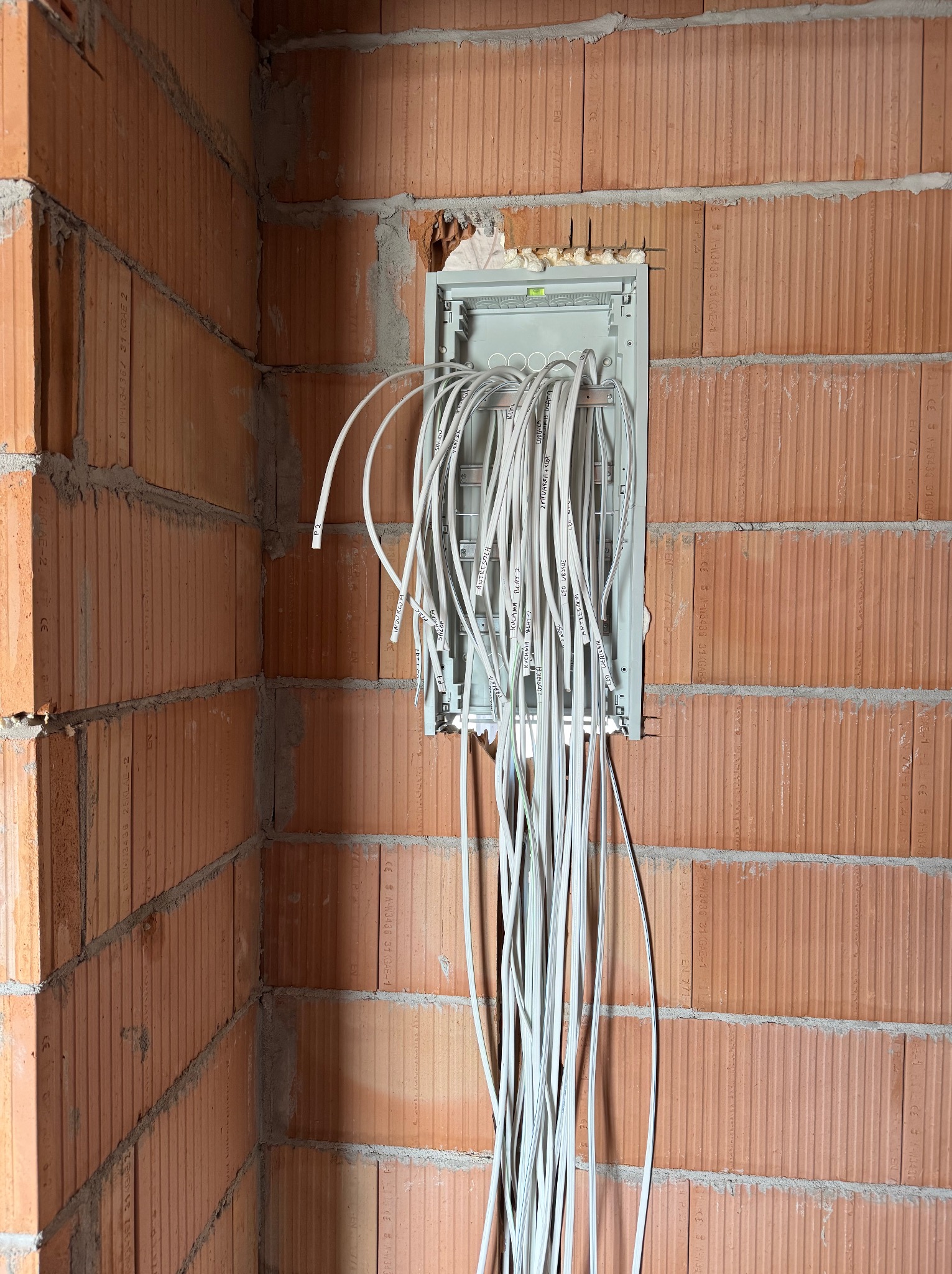 Rozdzielnica elektryczna z poprowadzonymi kablami w ścianie z czerwonej cegły, widoczne etykiety na przewodach opisujące ich przeznaczenie w nowym budynku.