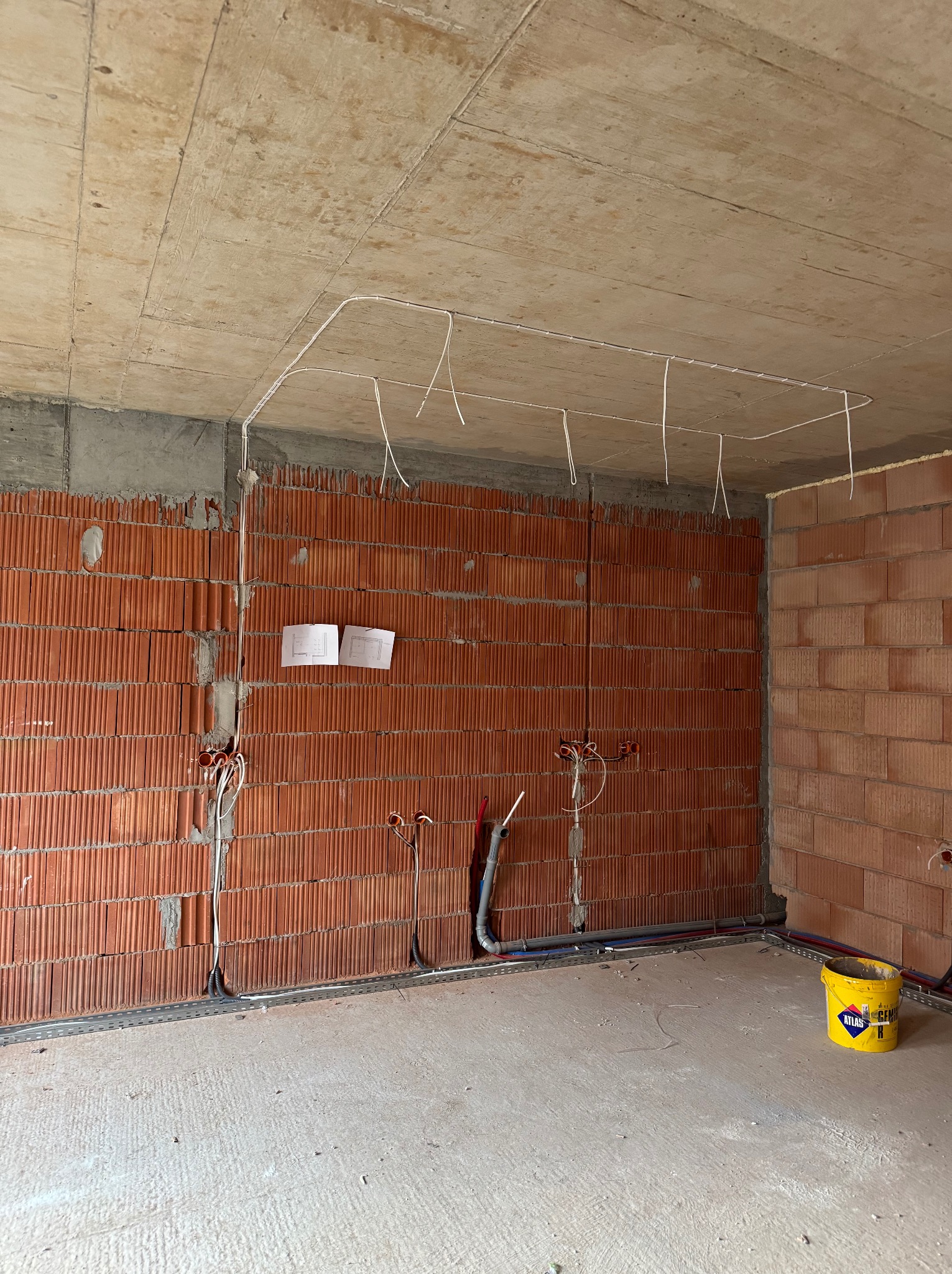 Surowy stan wnętrza z instalacją elektryczną: przewody poprowadzone pod sufitem, puszki elektryczne w ścianie z cegły dziurawki, rysunki techniczne na ścianie, widoczne rury instalacji wodnej...