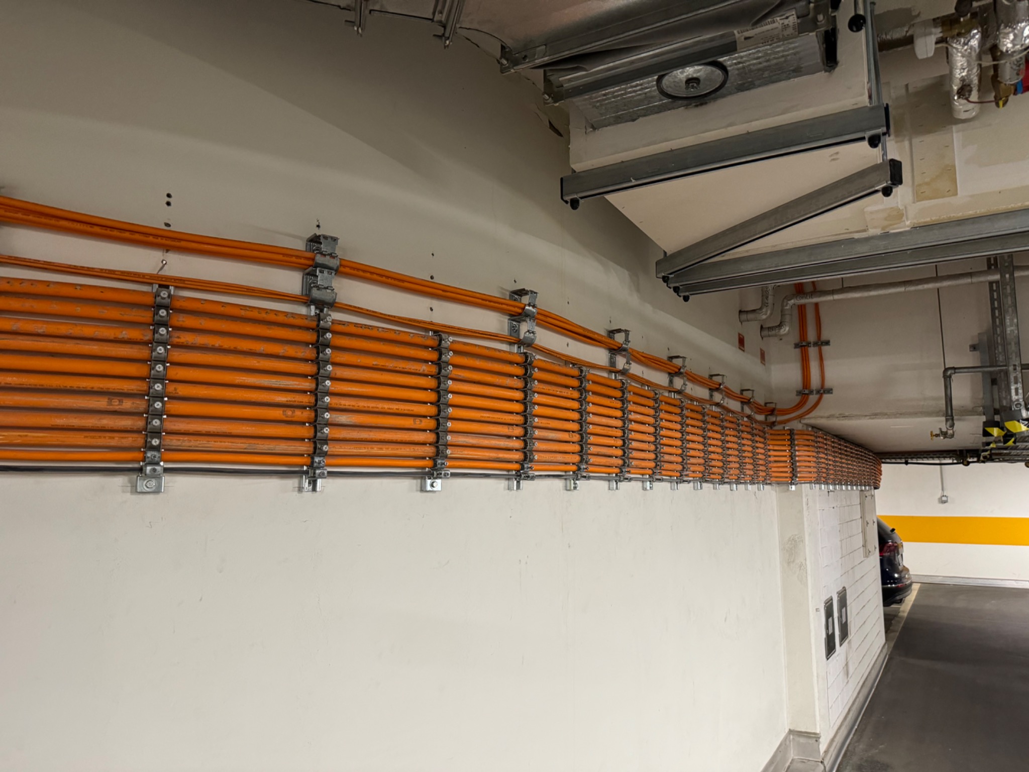 Równolegle ułożone pomarańczowe kable elektryczne przymocowane metalowymi uchwytami do ściany w garażu podziemnym, widoczne elementy infrastruktury nad głową, w tle zaparkowany samochód i żółty pas...