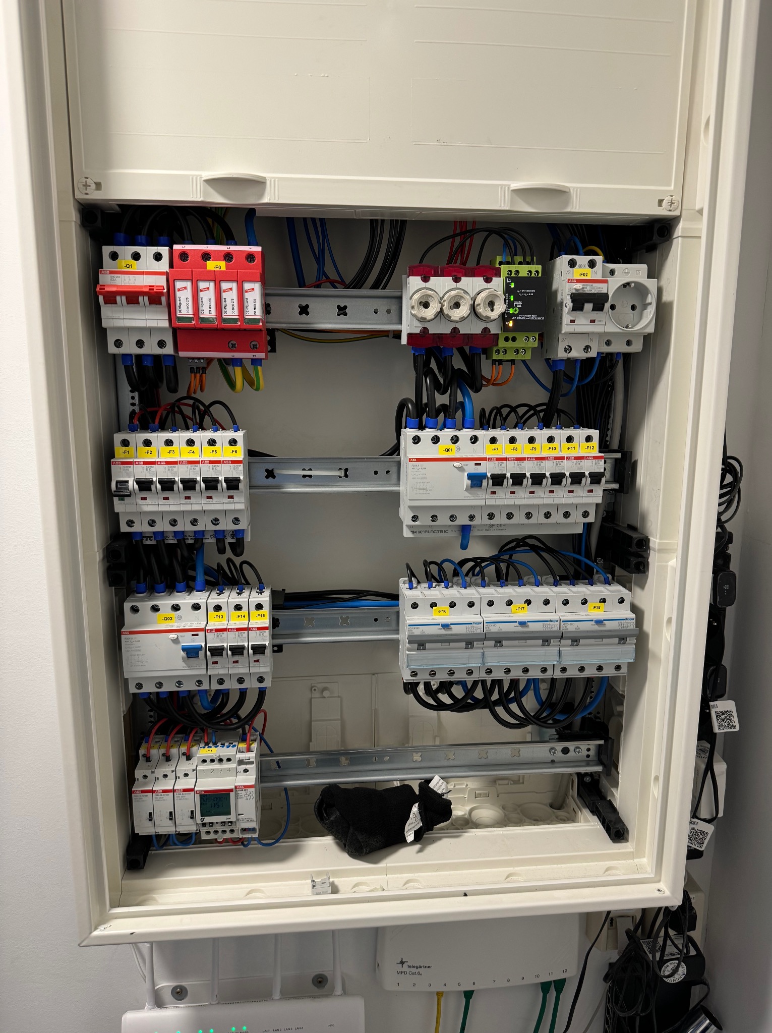 Otwarta skrzynka rozdzielcza z zainstalowanymi bezpiecznikami, wyłącznikami różnicowoprądowymi i zabezpieczeniami przeciwprzepięciowymi. Widoczne przewody w kolorach niebieskim, czarnym, brązowym...