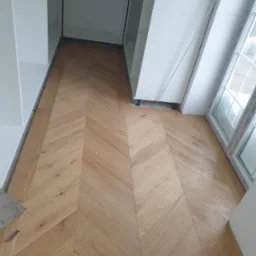 Ułożona parkietem jodełką podłoga z jasnego drewna w trakcie montażu szafy wnękowej i po montażu okna. Widoczne fragmenty białych mebli i okna PCV. Na podłodze leżą arkusze papieru ściernego.