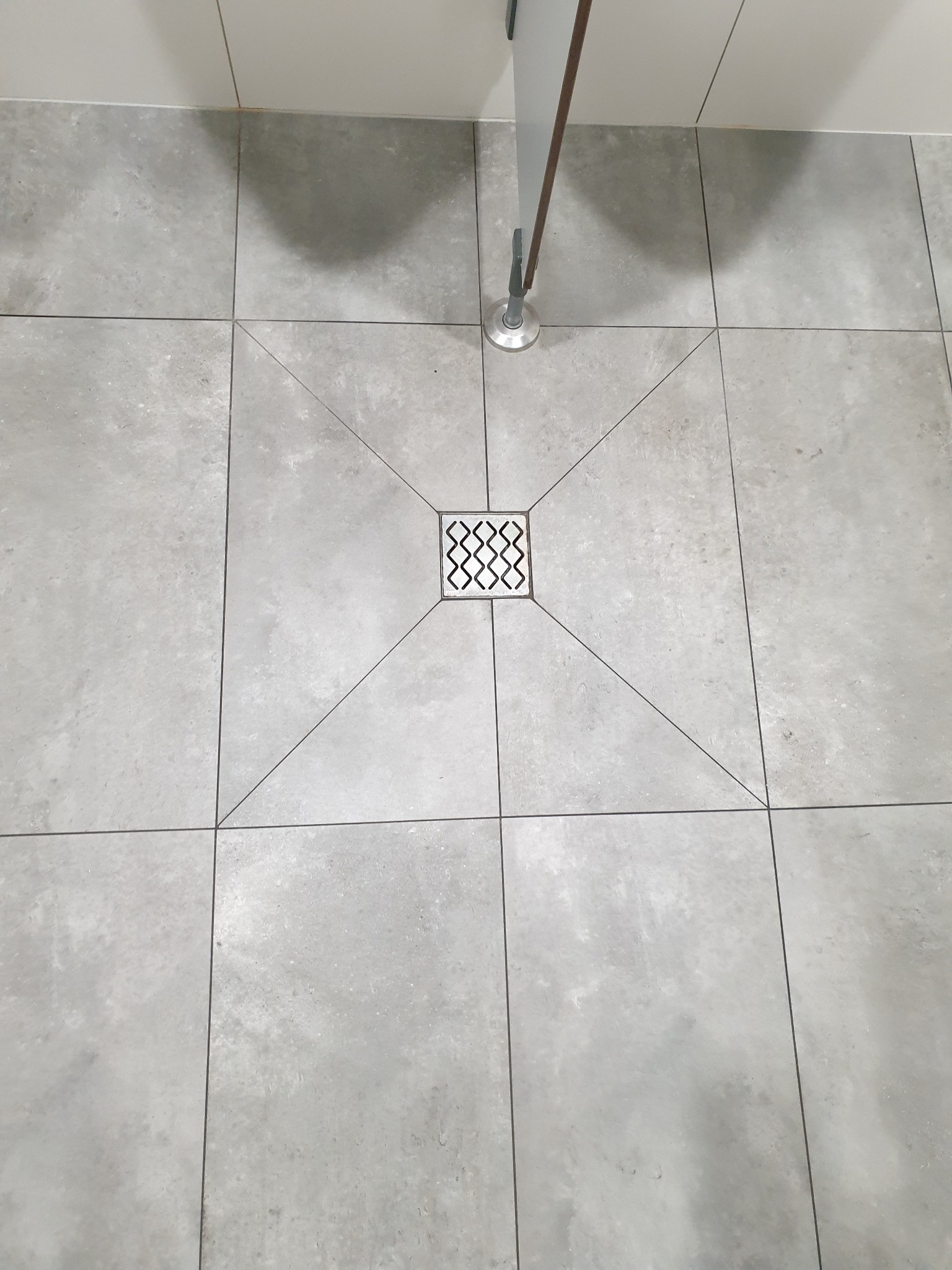 Szare, matowe płytki podłogowe w łazience z centralnie umieszczonym odpływem liniowym i widoczną nóżką meblową w tle.