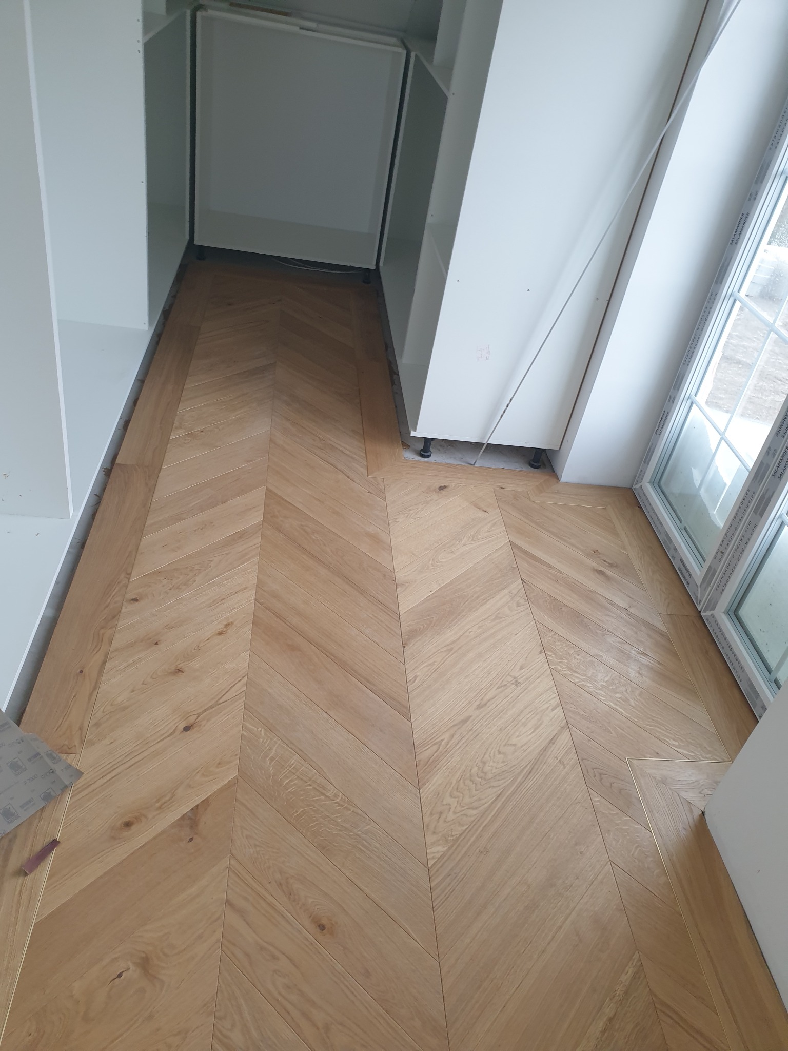 Ułożona parkietem jodełką podłoga z jasnego drewna w trakcie montażu szafy wnękowej i po montażu okna. Widoczne fragmenty białych mebli i okna PCV. Na podłodze leżą arkusze papieru ściernego.