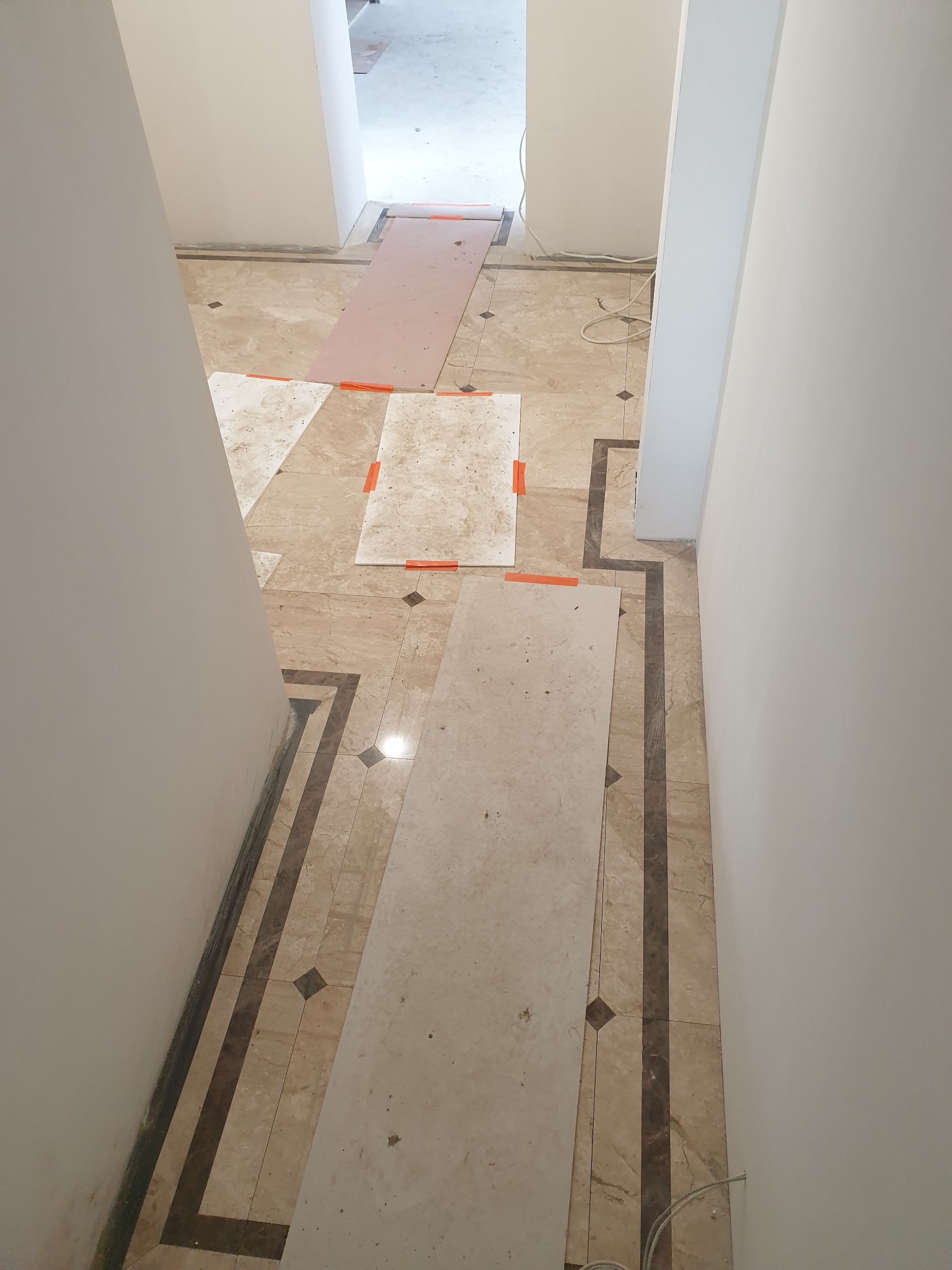 Korytarz w trakcie remontu z zabezpieczoną podłogą z płytek imitujących marmur. Na podłodze leżą płyty ochronne z pomarańczową taśmą zabezpieczającą. Widoczne białe ściany i kable elektryczne.