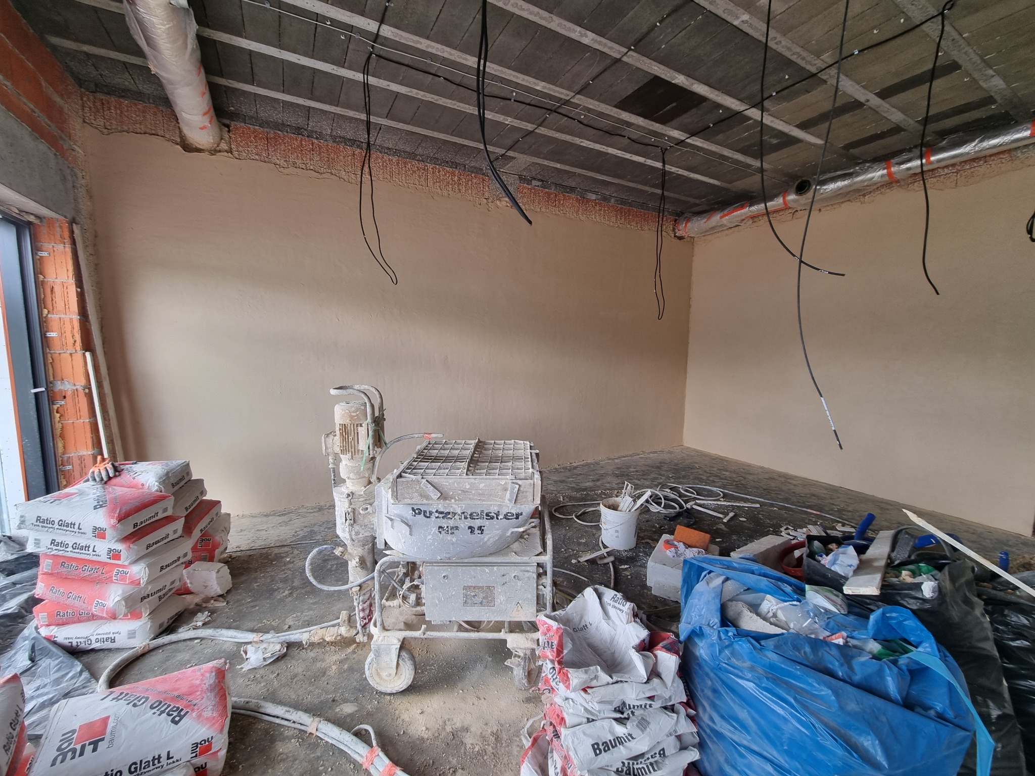 Wnętrze w stanie surowym po tynkowaniu maszynowym, widoczna maszyna Putzmeister MP 25, stosy worków z tynkiem gipsowym Ratio Glatt L i Baumin, instalacje pod sufitem.