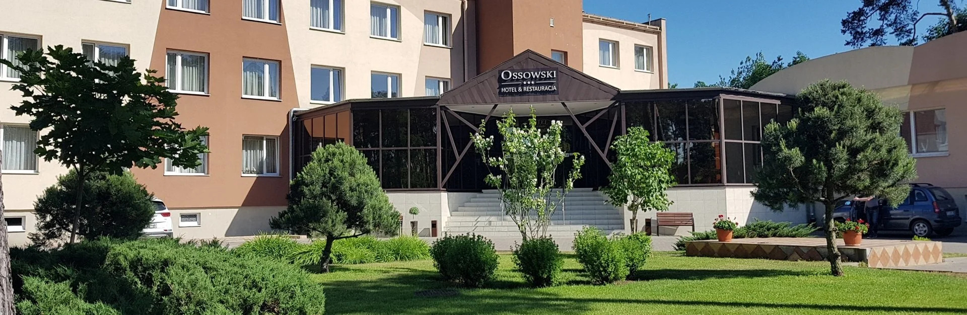 Wejście do hotelu Ossowski z ogrodem, widoczny budynek w odcieniach beżu i brązu, przeszklone wejście z ciemnobrązową konstrukcją, schody, zadbany trawnik z krzewami i drzewami, zaparkowane...