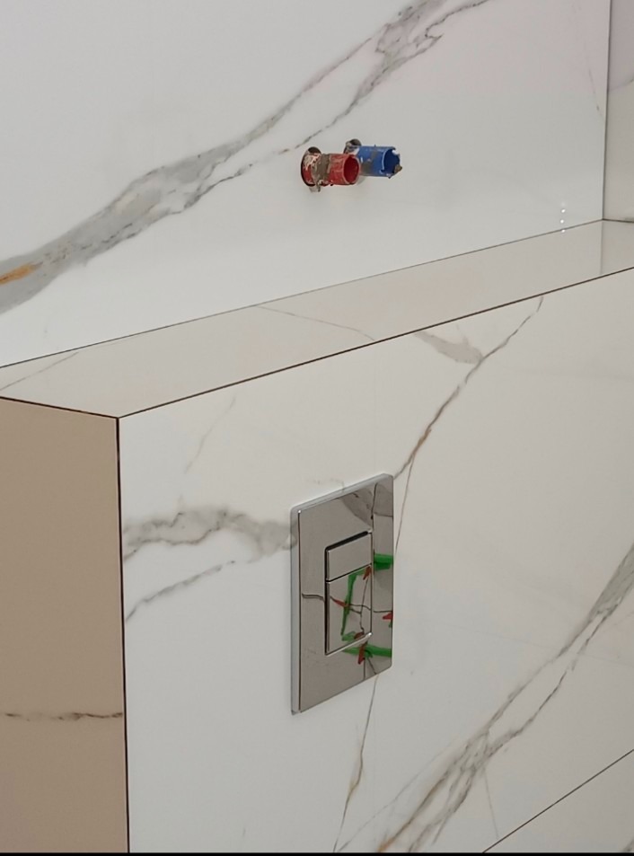 Ściana wyłożona białymi płytkami imitującymi marmur z zamontowanym chromowanym przyciskiem spłuczki i wystającymi rurami instalacji wodnej z zaślepkami