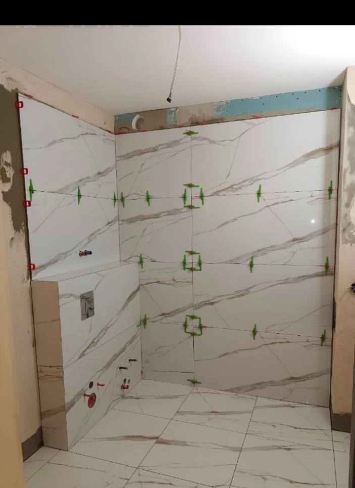 Łazienka w trakcie remontu: białe płytki imitujące marmur na ścianach i podłodze, zielone krzyżyki dystansowe, widoczne niedokończone prace przy suficie i instalacji wodnej.