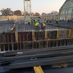 MW TRAST SPÓŁKA Z OGRANICZONĄ ODPOWIEDZIALNOŚCIĄ - Budowa fundamentów: robotnicy w kamizelkach odblaskowych i kaskach układają zbrojenie na placu budowy z widocznym dźwigiem i budynkami w tle.