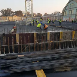 Budowa fundamentów: robotnicy w kamizelkach odblaskowych układają zbrojenie na placu budowy z widocznym żurawiem i budynkami w tle.