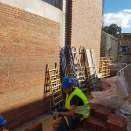 MW TRAST SPÓŁKA Z OGRANICZONĄ ODPOWIEDZIALNOŚCIĄ - Murarz w żółtej kamizelce i niebieskim kasku siedzi na palecie z cegłami na placu budowy, w tle ściana z czerwonej cegły i sprzęt budowlany.