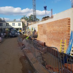 MW TRAST SPÓŁKA Z OGRANICZONĄ ODPOWIEDZIALNOŚCIĄ - Budowa ściany z cegły z użyciem dźwigu i betoniarki, widoczni pracownicy w kaskach i kamizelkach odblaskowych, na pierwszym planie barierki ochronne i porozrzucane cegły.