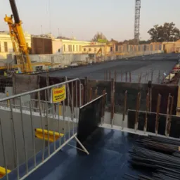 Widok placu budowy z żurawiami, zbrojeniami i tabliczką ostrzegawczą 'Uwaga! Głębokie wykopy'.