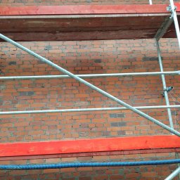 MW TRAST SPÓŁKA Z OGRANICZONĄ ODPOWIEDZIALNOŚCIĄ - Ceglana ściana z szarymi rusztowaniami i czerwonymi deskami, widok z dołu, niebieski przewód w dolnej części kadru.