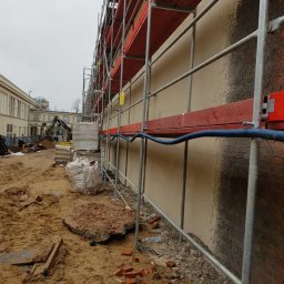 MW TRAST SPÓŁKA Z OGRANICZONĄ ODPOWIEDZIALNOŚCIĄ - Elewacja budynku w trakcie renowacji z widocznymi rusztowaniami, stertą piasku, gruzem i koparką w tle.