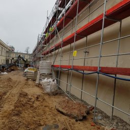 MW TRAST SPÓŁKA Z OGRANICZONĄ ODPOWIEDZIALNOŚCIĄ - Ściana budynku w trakcie prac, zabezpieczona rusztowaniem z czerwonymi deskami, w tle koparka i materiały budowlane, teren wokół pokryty piaskiem i gruzem.