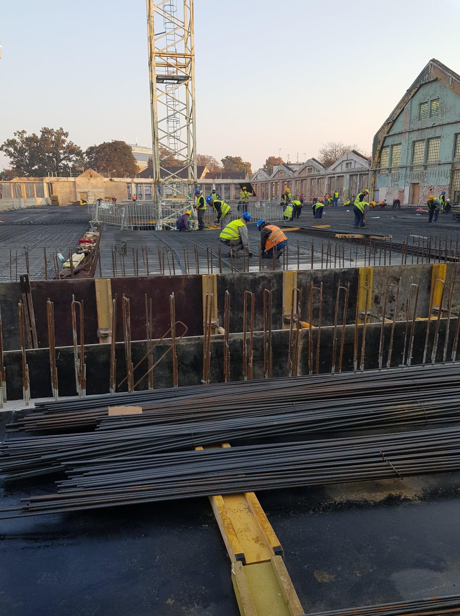 Budowa fundamentów: robotnicy w kamizelkach odblaskowych i kaskach układają zbrojenie na placu budowy z widocznym dźwigiem i budynkami w tle.