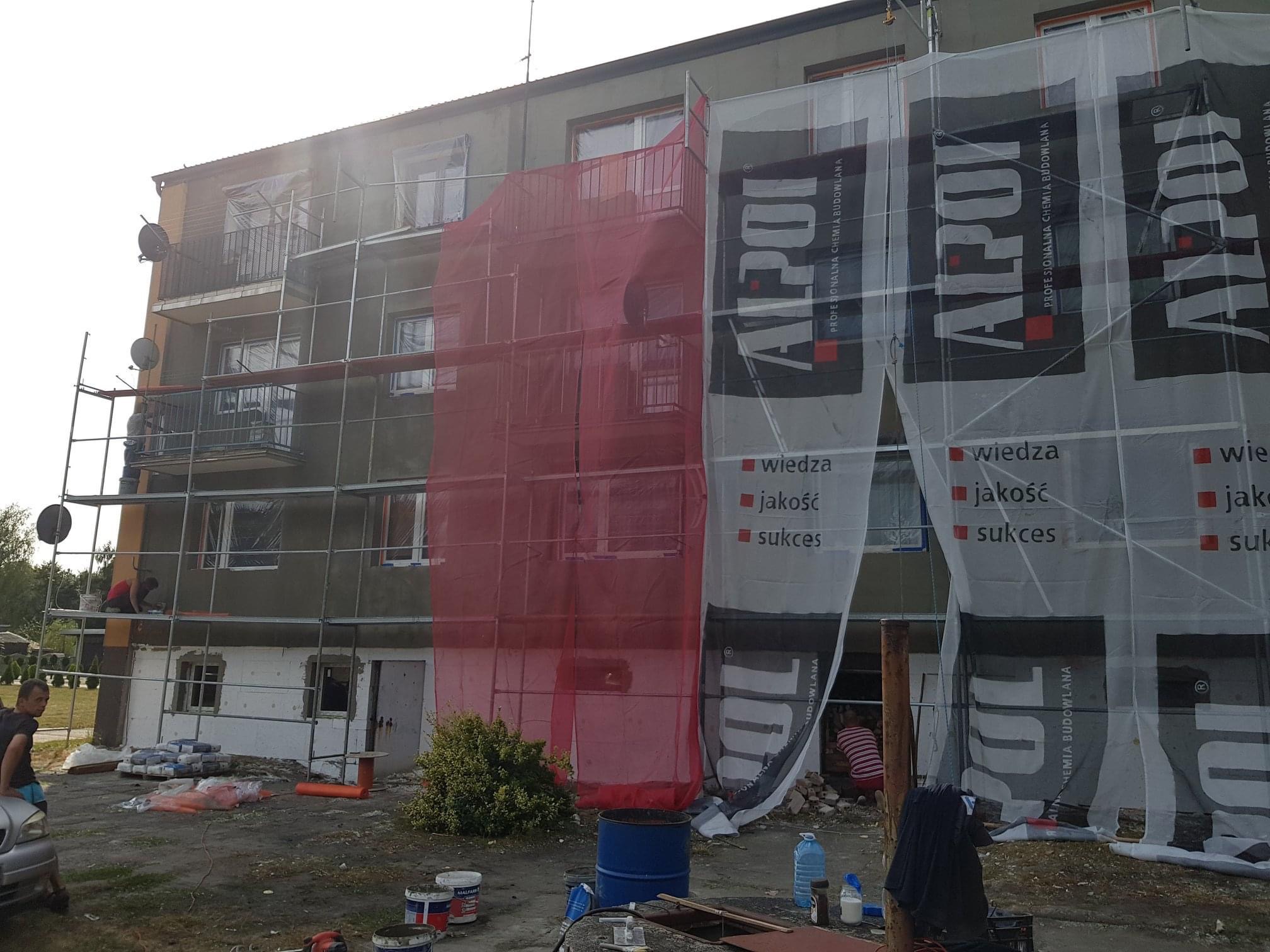 Budynek wielorodzinny w trakcie prac elewacyjnych z użyciem rusztowania i siatki ochronnej, widoczne materiały budowlane i reklama firmy Alpol.