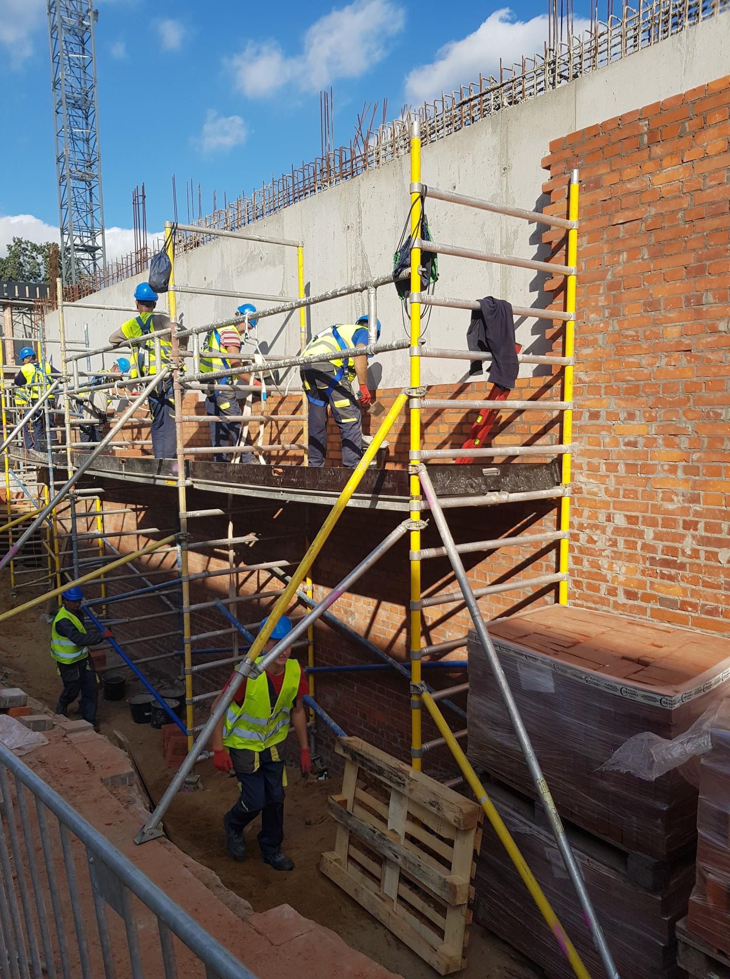 Murowanie ściany z czerwonej cegły na rusztowaniu; widoczni robotnicy w kaskach i kamizelkach odblaskowych, palety z cegłami w folii, betonowy mur w tle.