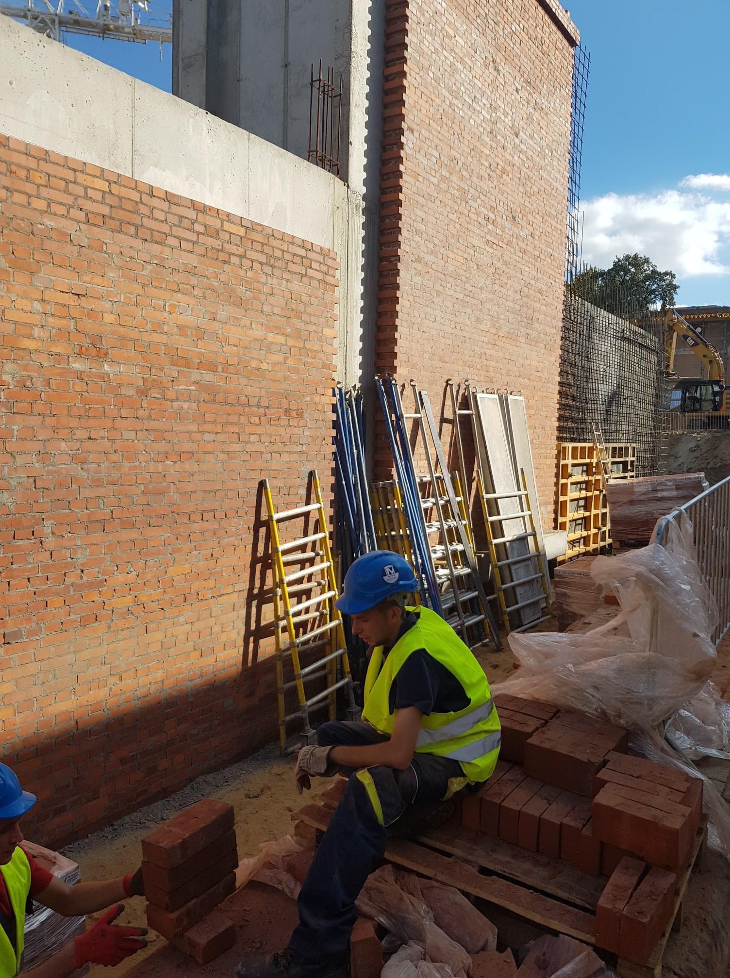 Murarz w żółtej kamizelce i niebieskim kasku siedzi na palecie z cegłami na placu budowy, w tle ściana z czerwonej cegły i sprzęt budowlany.