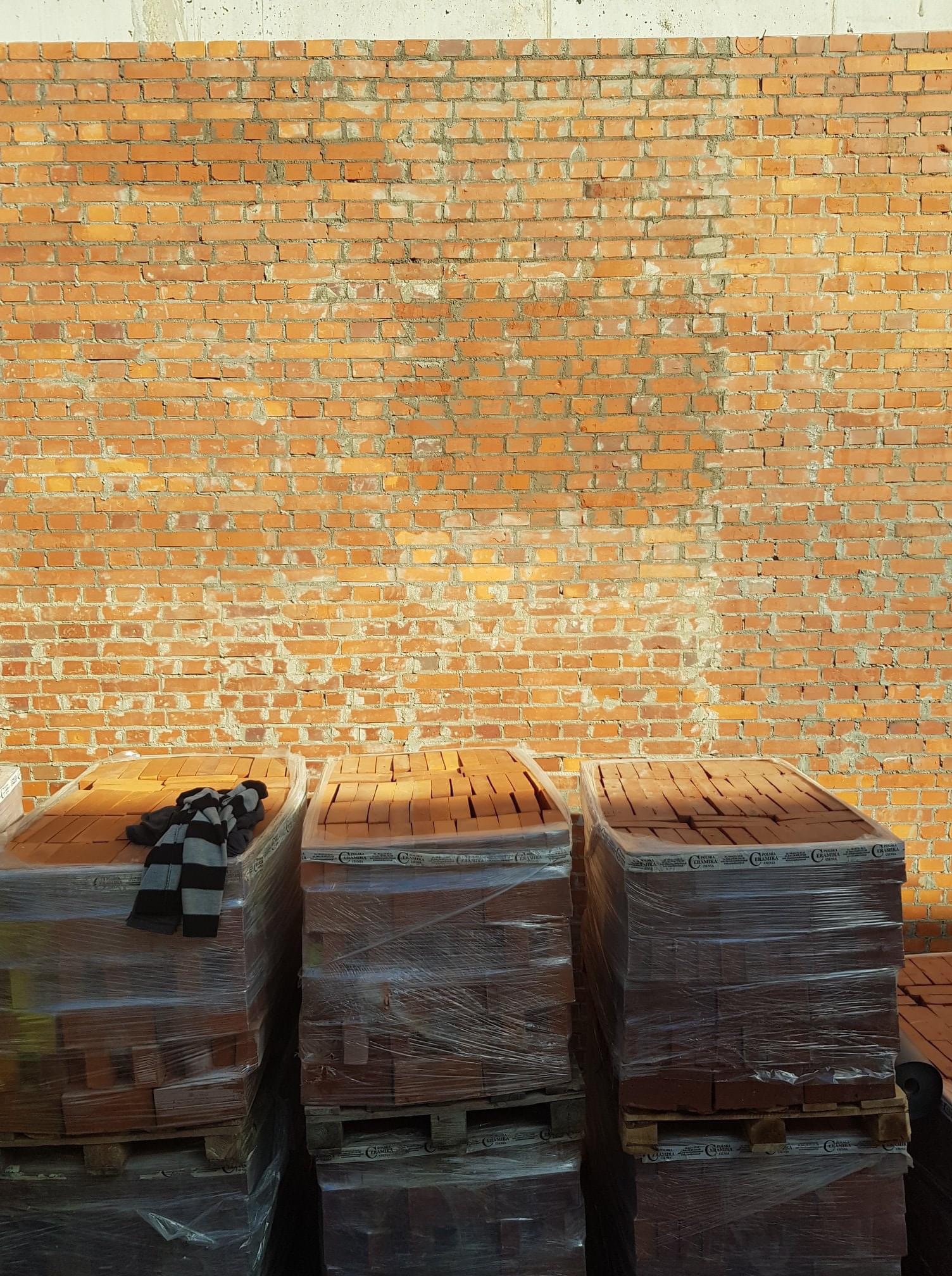 Palety z czerwoną cegłą ceramiczną, zapakowane w folię, stoją na tle muru z cegły w Łochowie. Na jednej z palet leży czapka w czarno-szare paski.