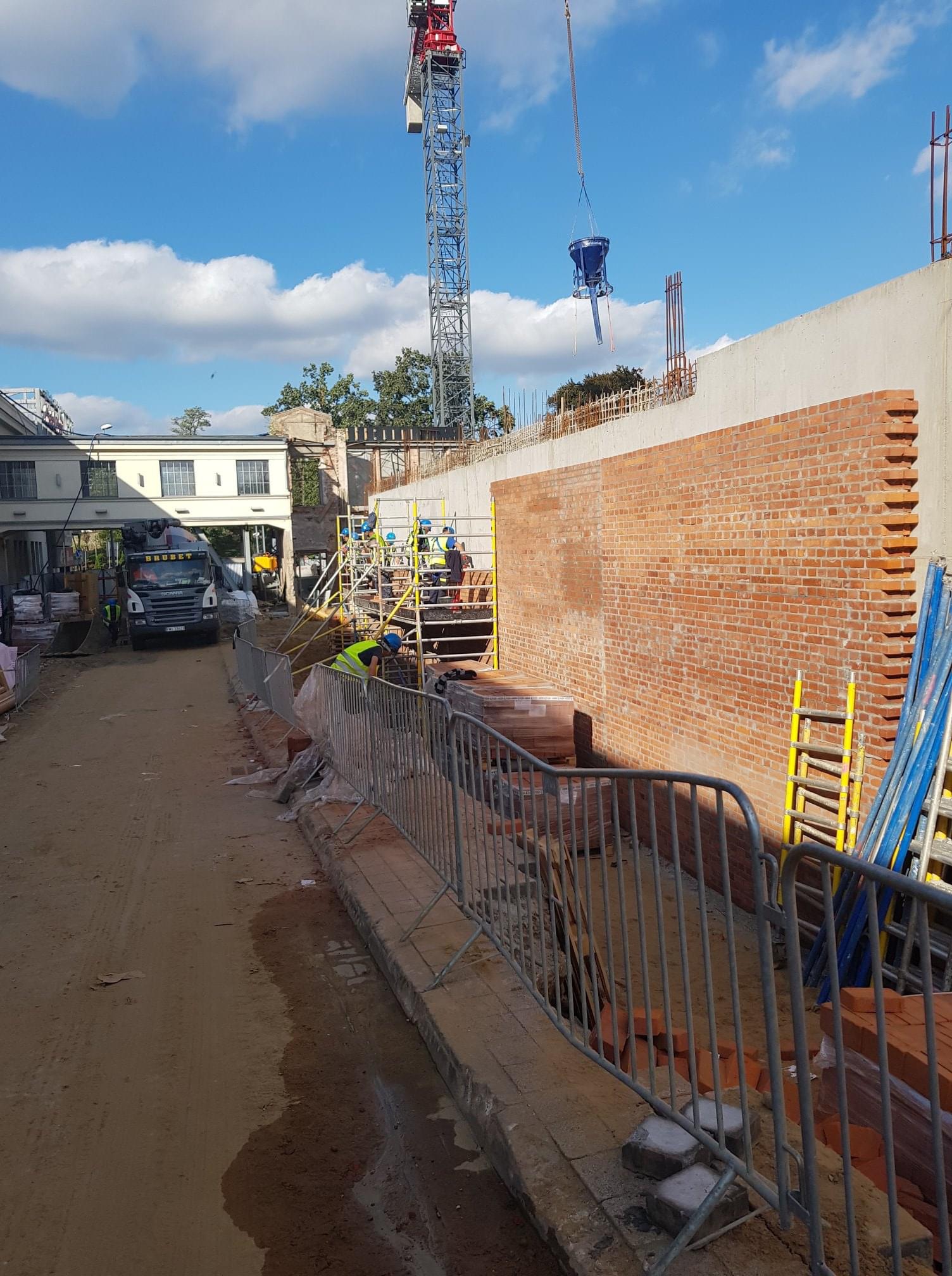 Budowa ściany z cegły z użyciem dźwigu i betoniarki, widoczni pracownicy w kaskach i kamizelkach odblaskowych, na pierwszym planie barierki ochronne i porozrzucane cegły.