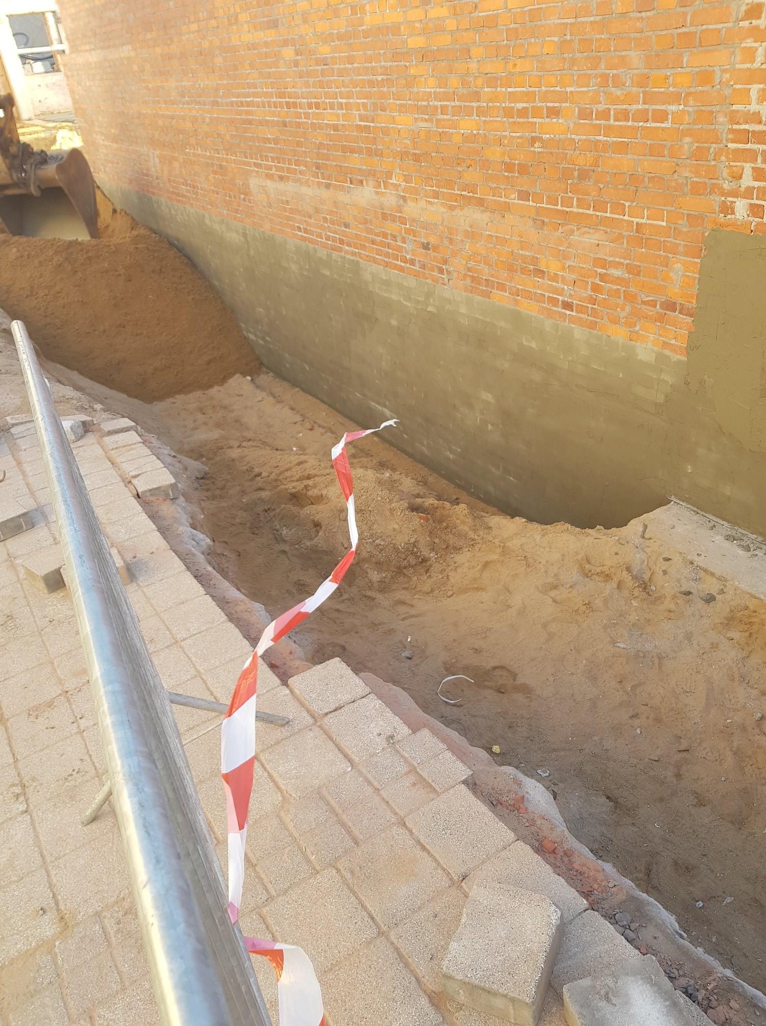 Wykop fundamentów przy murowanym budynku z zabezpieczoną hydroizolacją, widoczny fragment koparki, taśma ostrzegawcza i chodnik z kostki brukowej.