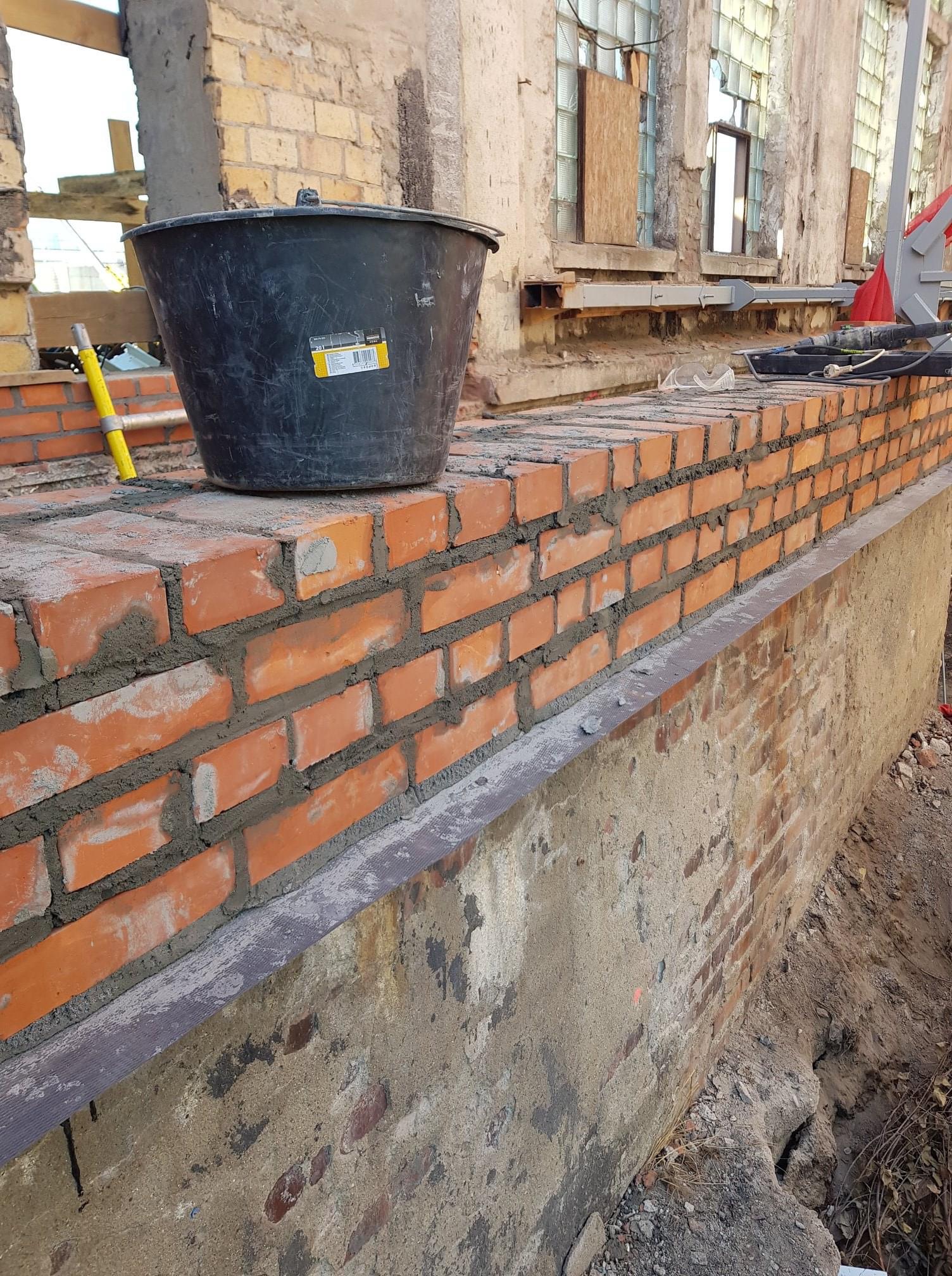 Wznoszenie muru z czerwonej cegły, z widocznym czarnym wiadrem na górze i żółtą poziomicą, na tle budynku z oknami częściowo zabezpieczonymi płytami.