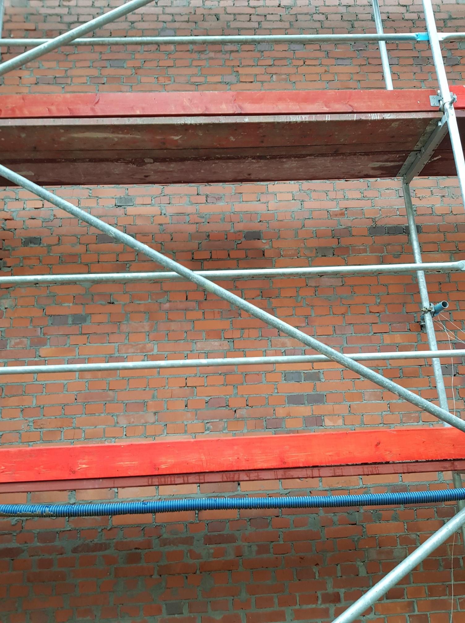 Ceglana ściana z szarymi rusztowaniami i czerwonymi deskami, widok z dołu, niebieski przewód w dolnej części kadru.
