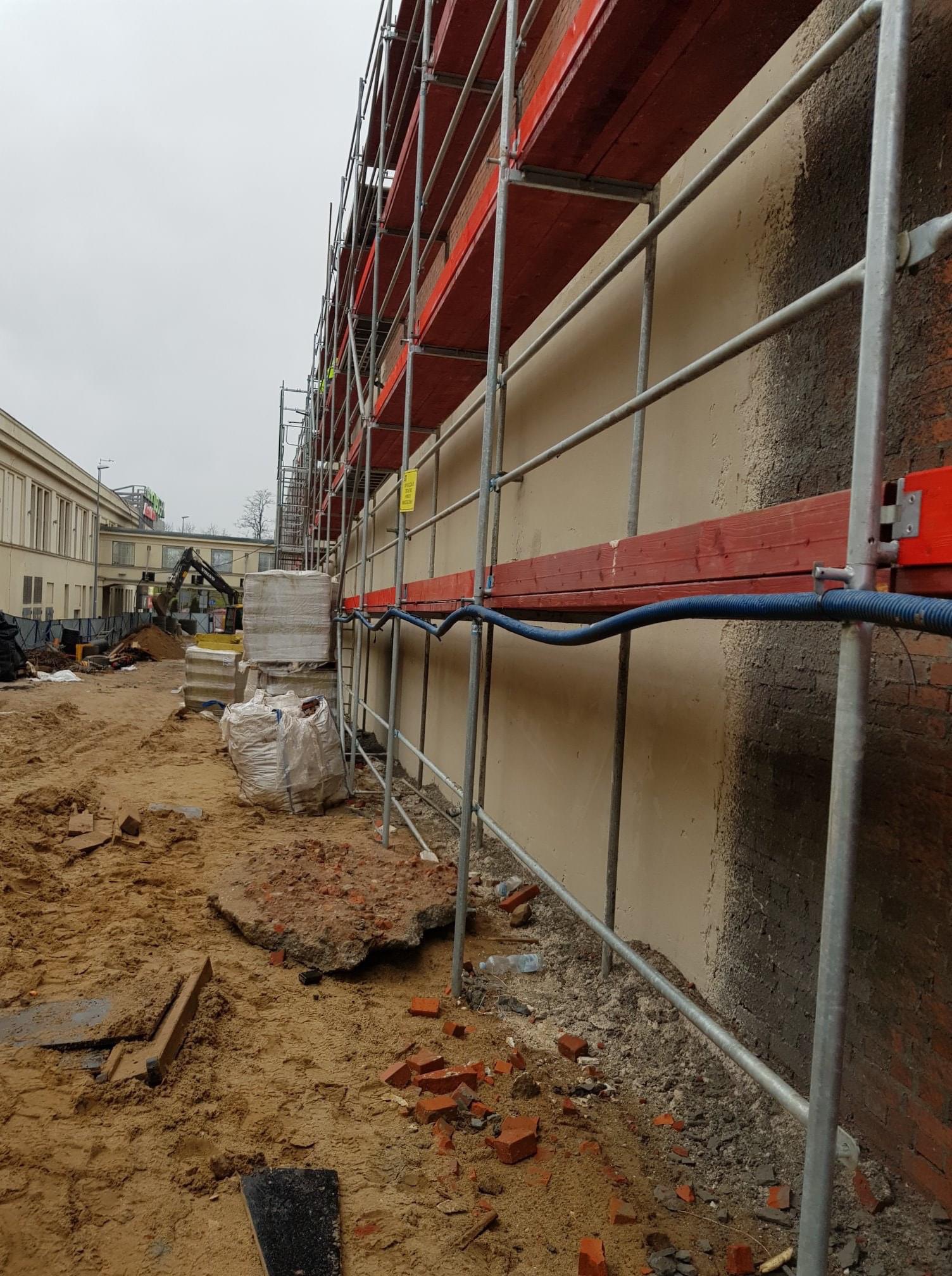 Elewacja budynku w trakcie renowacji z widocznymi rusztowaniami, stertą piasku, gruzem i koparką w tle.