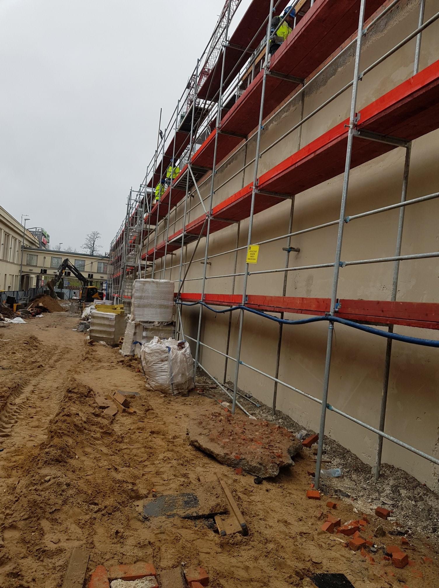 Ściana budynku w trakcie prac, zabezpieczona rusztowaniem z czerwonymi deskami, w tle koparka i materiały budowlane, teren wokół pokryty piaskiem i gruzem.