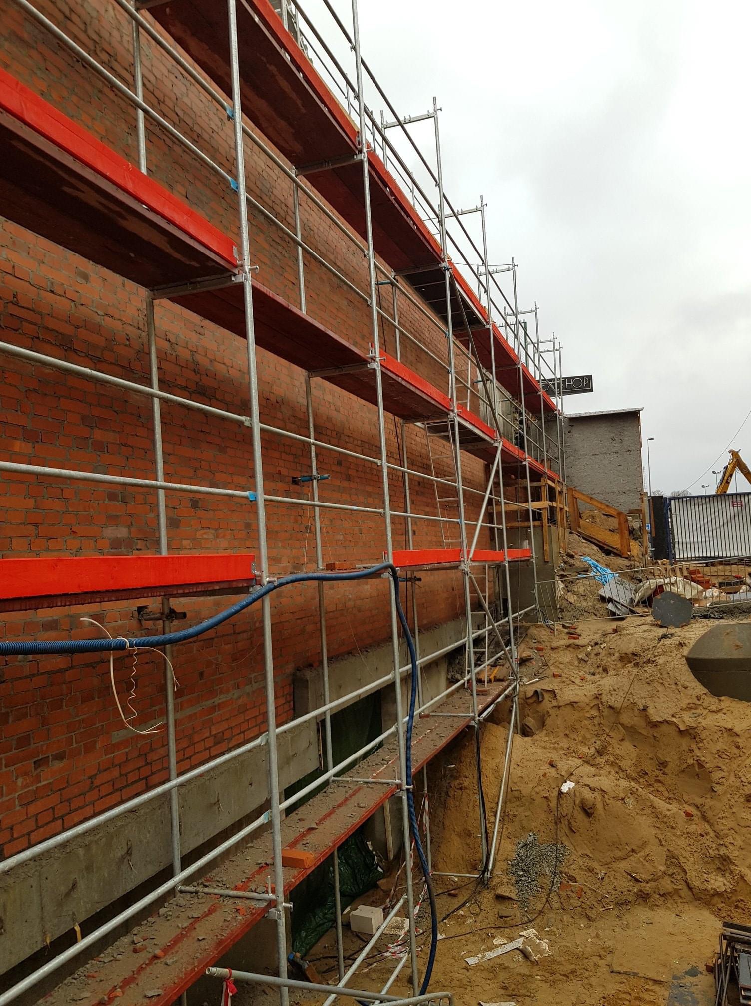 Budynek z czerwonej cegły w trakcie budowy, otoczony rusztowaniem z czerwonymi deskami, widoczny wykop w ziemi i elementy konstrukcyjne.