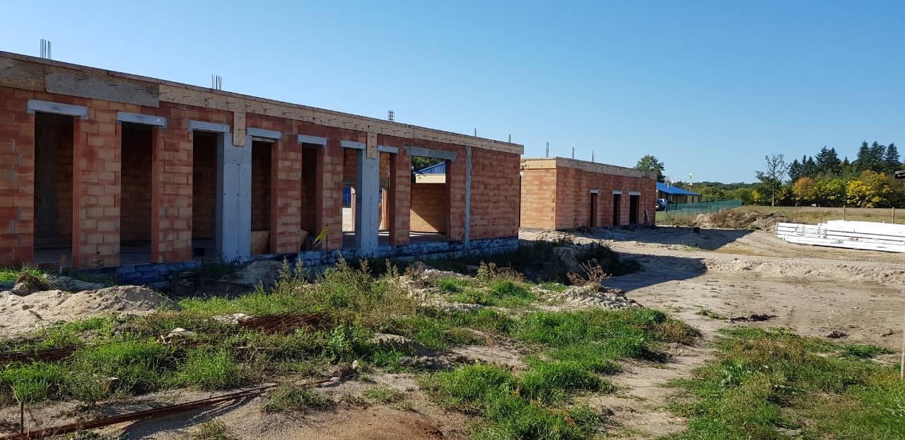 Wznoszenie ścian parterowych budynków z czerwonej cegły, widoczne betonowe nadproża okien i drzwi, teren budowy z piaskiem i trawą, w tle zalesione wzgórze pod błękitnym niebem.