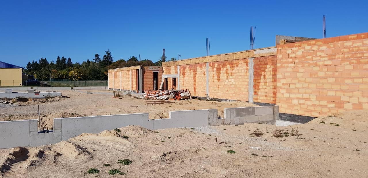 Wznoszenie ścian z czerwonej cegły na placu budowy, widoczne fundamenty i zbrojenia, w tle drzewa pod błękitnym niebem.