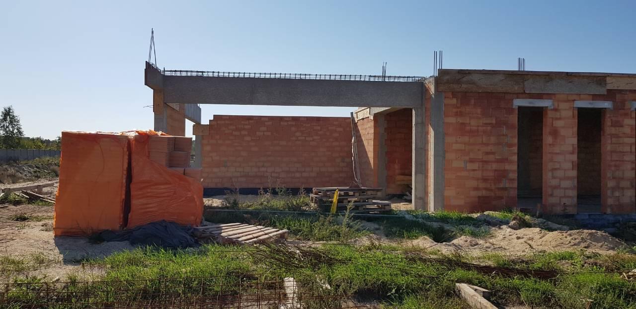 Wznoszenie domu murowanego z czerwonej cegły, widoczne zbrojenia stropu i stos cegieł zabezpieczony pomarańczową folią