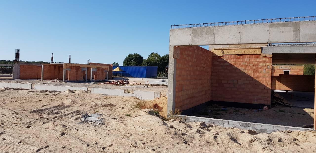 Wznoszenie ścian z czerwonej cegły na fundamencie betonowym, widoczne zbrojenia na górze ścian, piasek na placu budowy, niebieski kontener w tle.