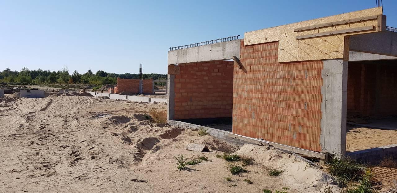 Wznoszenie ścian parterowego domu z czerwonej cegły ceramicznej, widoczne betonowe słupy i fundamenty, teren budowy piaszczysty z widocznymi śladami opon.