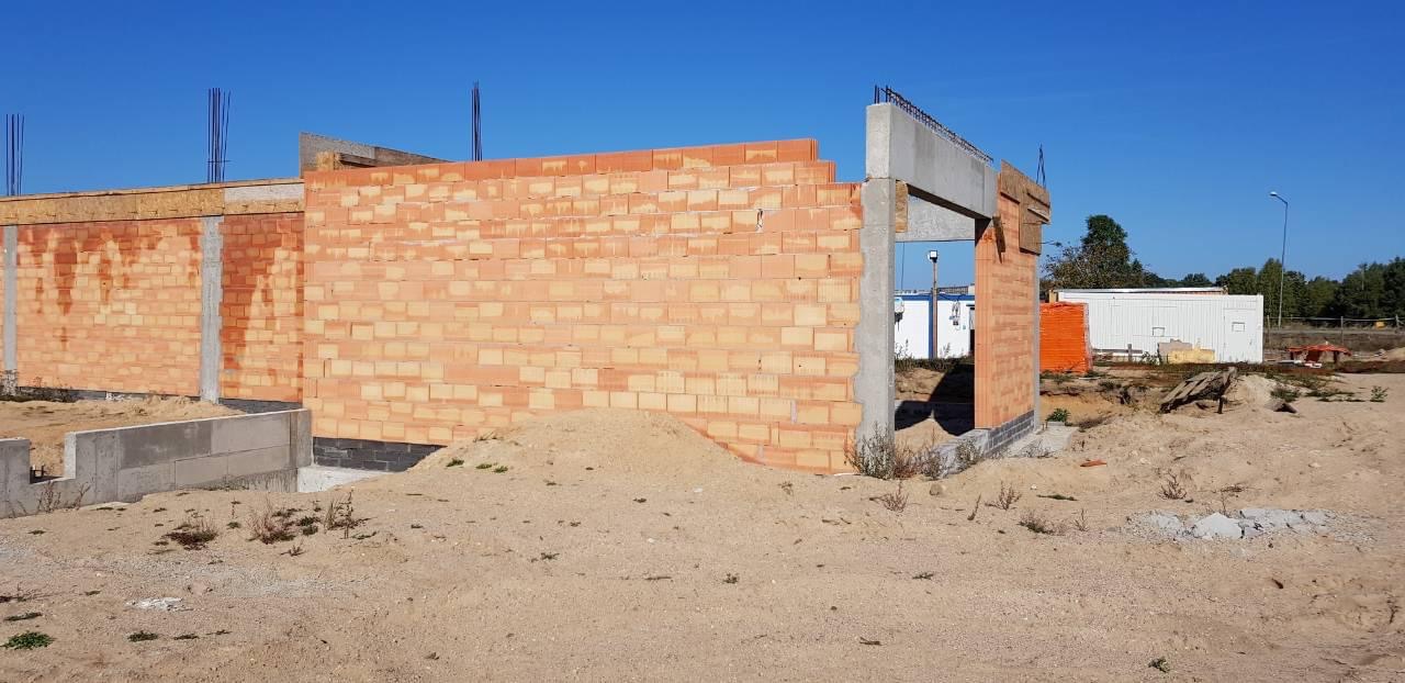 Wznoszone ściany z bloczków ceramicznych na budowie domu jednorodzinnego, widoczne zbrojenia i betonowe nadproża, w tle baraki budowlane.
