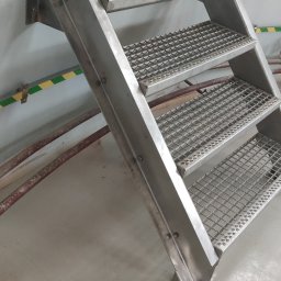 Mateusz Mazurek - Trzy stopnie metalowe z kratownicą, przymocowane do ściany, z widocznymi śrubami mocującymi i rurami w tle.