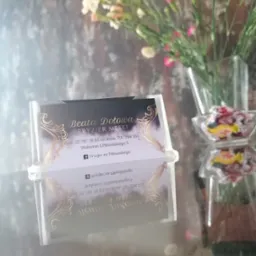 Wizytówka salonu z imieniem Beata Dołowa, specjalizującego się we fryzjerstwie męskim, umieszczona w przezroczystym stojaku, na tle wazonu z goździkami i szklanej miseczki z cukierkami.