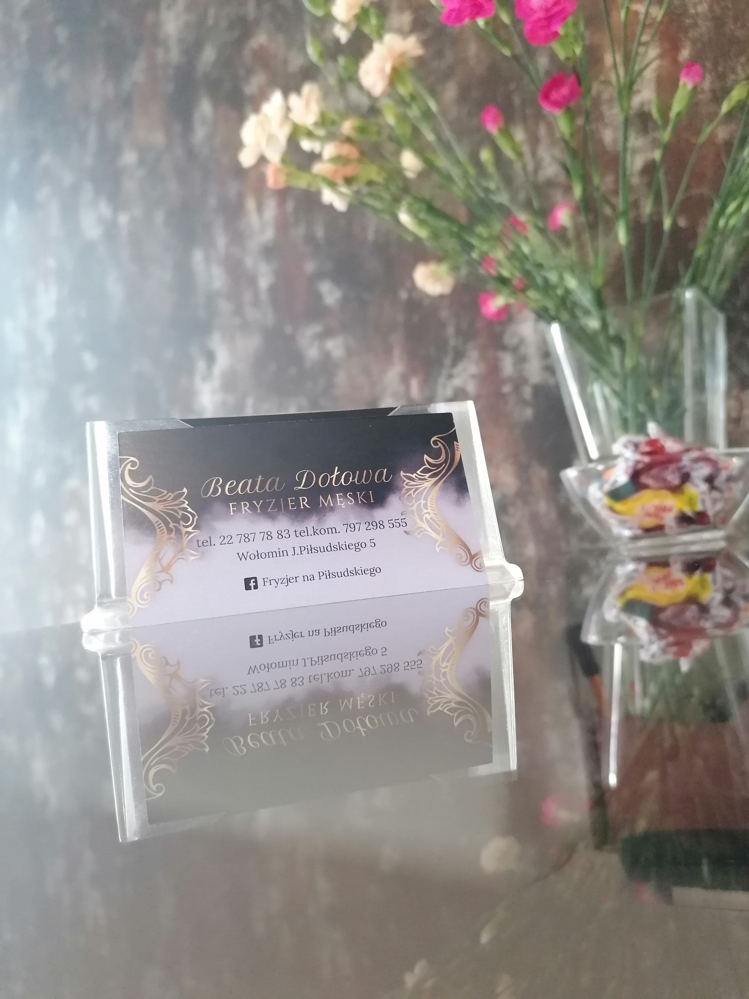 Wizytówka salonu z imieniem Beata Dołowa, specjalizującego się we fryzjerstwie męskim, umieszczona w przezroczystym stojaku, na tle wazonu z goździkami i szklanej miseczki z cukierkami.