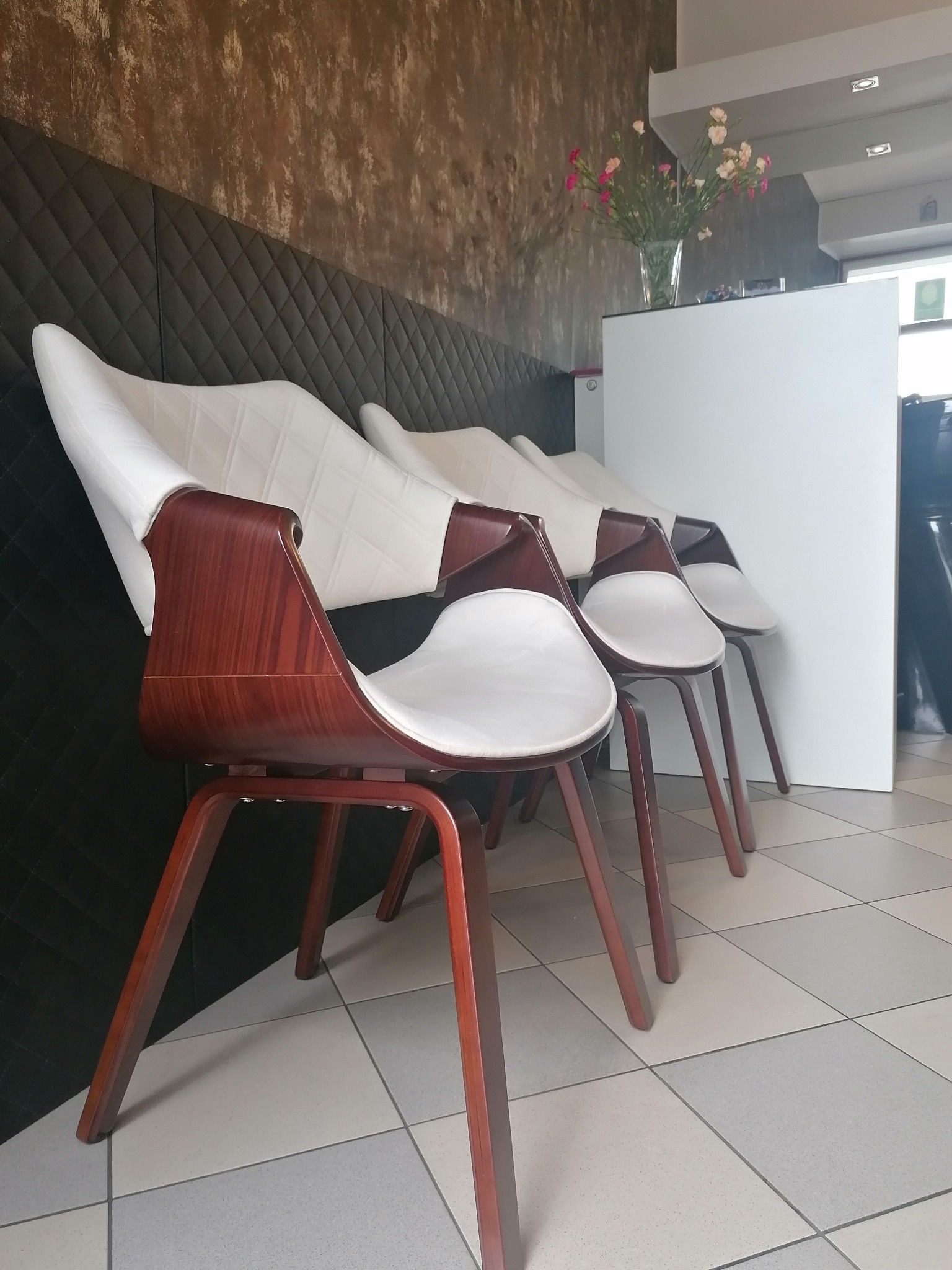 Czekając na wizytę: Rząd nowoczesnych krzeseł z drewnianymi ramami i białymi siedziskami w salonie, w tle dekoracyjna ściana i wazon z kwiatami na białej recepcji.