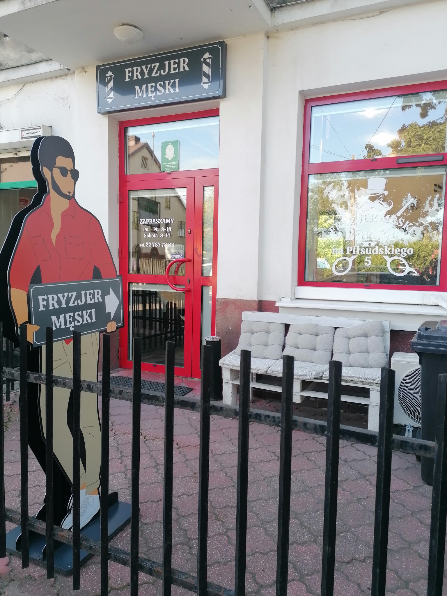 Fasada salonu z szyldem 'Fryzjer Męski', przed wejściem stoi kartonowa postać mężczyzny w okularach słonecznych trzymająca tabliczkę z napisem 'Fryzjer Męski', w oknie naklejka z logo salonu.