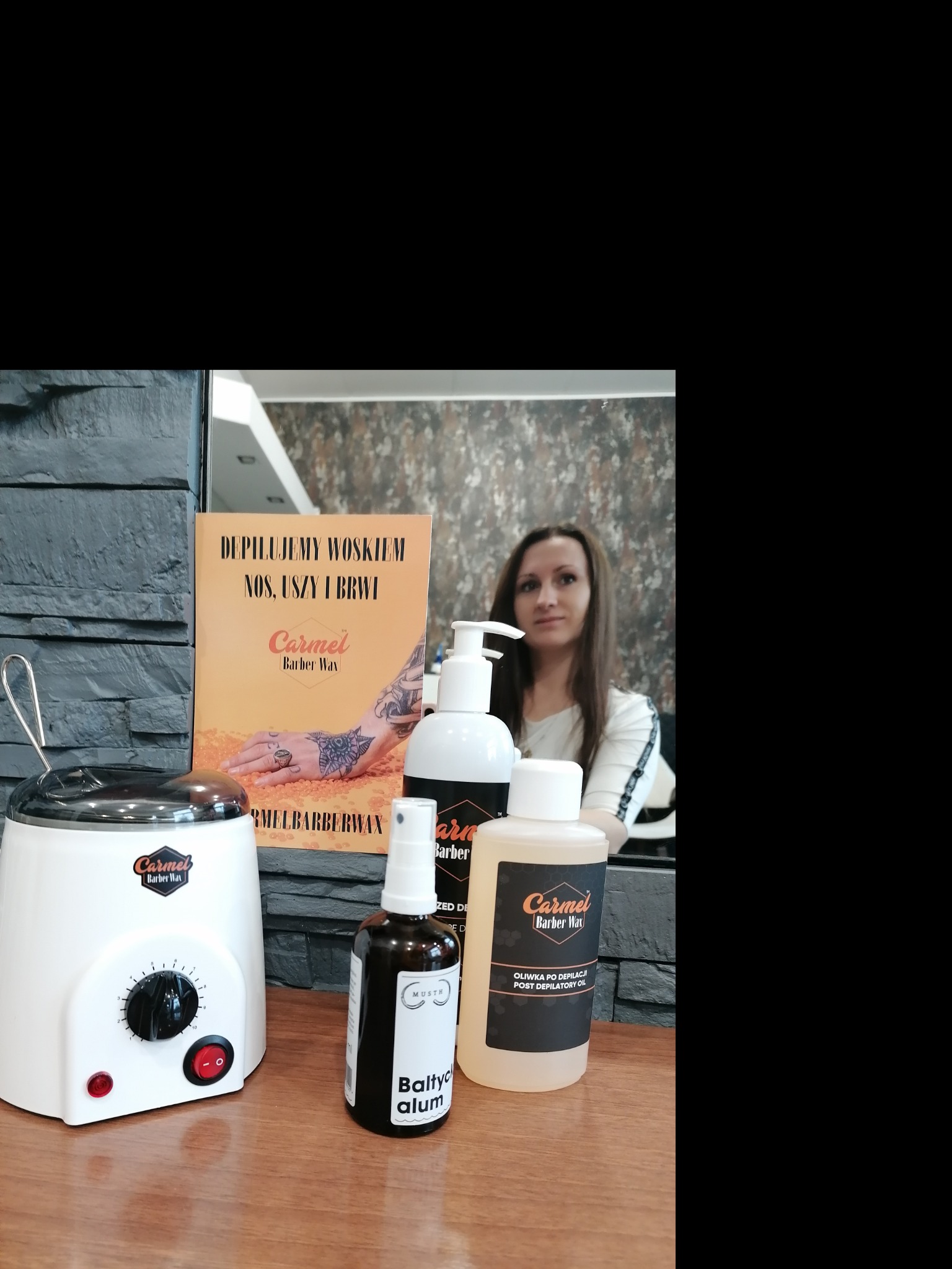 Zestaw produktów do depilacji woskiem marki Carmel Barber Wax, w tym podgrzewacz do wosku, oliwka po depilacji i spray, na drewnianym blacie, z odbiciem kobiety w lustrze w tle.