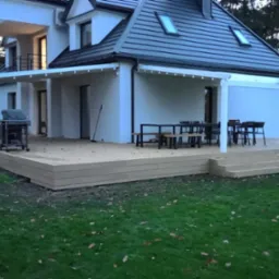 Rozległy taras z desek kompozytowych z pergolą przy nowoczesnym domu, z widocznym grillem i meblami ogrodowymi, otoczony trawnikiem z opadłymi liśćmi.