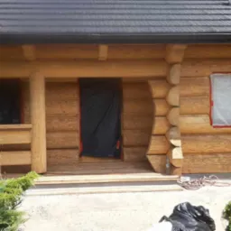 Dom z bali w trakcie budowy lub remontu, okna i drzwi zabezpieczone folią, widoczne elementy drewniane i podmurówka