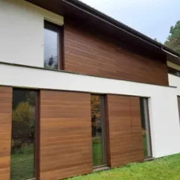 Nowoczesny dom z elewacją z paneli drewnopodobnych w odcieniach brązu, z dużymi oknami odbijającymi zieleń ogrodu.