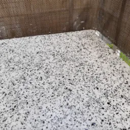 Biała powierzchnia z czarnymi i szarymi drobnymi elementami, wykończona srebrną listwą przy ścianie z dekoracyjnym panelem z brązowej plecionki, widoczny fragment zieleni.